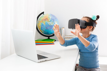 Little kid schoolgirl sit and study at white desk with pc laptop. School distance education at home during quarantine concept. Watching in headset, virtual glasses