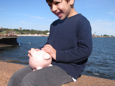 Money saving in piggy bank. A happy young boy slips a coin into a pink ceramic piggyback in his sunlit backyard.