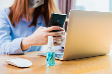 Asian young business woman wearing face mask protective using smart mobile phone for reading SMS message near laptop computer at home office quarantines disease coronavirus or COVID-19, work from home