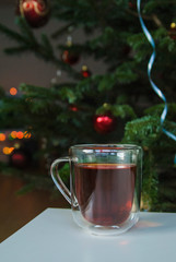 A glass cup of tea stands on a white table. In the background is a green Christmas tree decorated for New Year and Christmas.