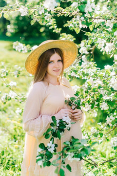 Beautiful pregnant woman wearing big hat and beige dress in blooming garden. Spring nature sunny day.