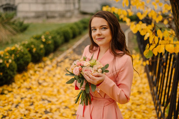 Naklejka premium Portrait of happy sensual young brunette bride in rose wedding dress and standing in autumn park near vintage cathedral and holding wedding bouquet.