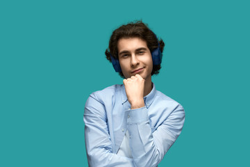 Fototapeta premium What do you say?! Portrait of a young beautiful man wearing white t-shirt and blue shirt in blue headphones. Thought holding a fist at his chin