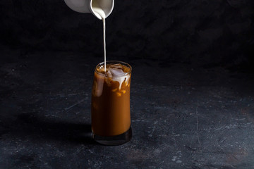 Ice coffee frappe in tall glass. Cool summer drink on a dark background in low key. A stream of milk pours into the coffee. Close up, copy space for text