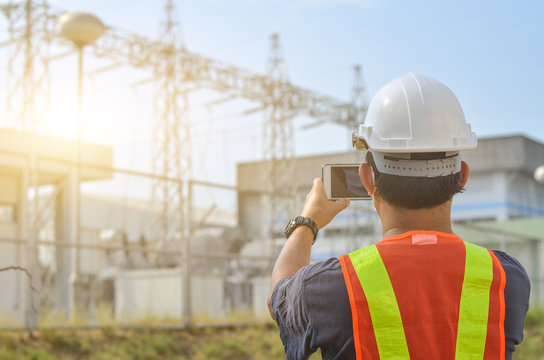 Engineers use mobile phones to photograph high-voltage electricity poles against sub-station background.