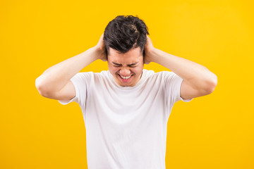Portrait Aaian handsome young man standing wearing white t-shirt he covering his ears with hands and shouting opened mouth shriek annoyed expression, studio shot isolated yellow background
