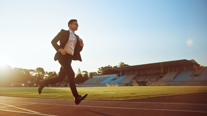 Young Serious Businessman in a Suit Running in an Outdoors Stadium. He Wears Glasses and is Holding...