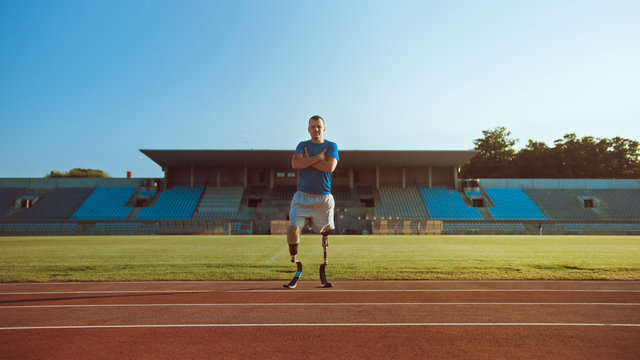 Athletic Disabled Fit Man with Prosthetic Running Blades is Posing with Crossed Arms on an Outdoor Stadium on a Sunny Afternoon. Amputee Runner Standing on a Track. Motivational Sports Shot.