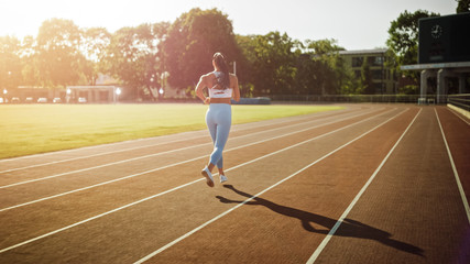 Fototapeta premium Beautiful Fitness Girl in Light Blue Athletic Top and Leggings Jogging in the Stadium. She is Running on a Warm Summer Afternoon. Athlete Doing Her Routine Sports Practice on a Track.