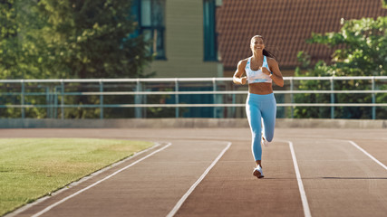 Obraz premium Beautiful Fitness Woman in Light Blue Athletic Top and Leggings Jogging in a Stadium. She is Running on a Warm Summer Afternoon. Athlete Doing Her Routine Sports Practice on a Track.