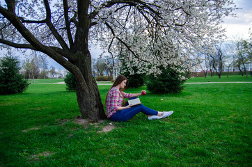 
Beautiful girl with long hair reads a book near a flowering tree. Plaid shirt, jeans and white sneakers