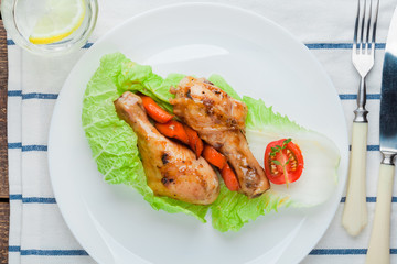 Fried chicken legs in caramelized sauce on a white plate with garnish of baked and fresh vegetables on a light tablecloth.  Glass of water, vintage cutlery. Mexican, Organic products. 