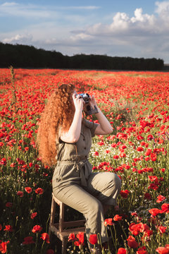 Young beautiful red-hear girl takes photos in the red poppy flowers field