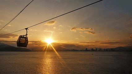 Cable way through the bay. The rays of the setting sun color the sky and sea in a golden color. The cable car cabin moves to the island. On the horizon are silhouettes of buildings and mountains. 