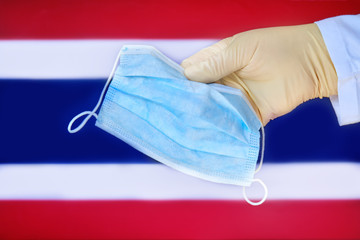 Medical mask in the hands of a doctor and the Thailand flag, copy space. Protective glove with mask close-up on the background of the state Thai symbol