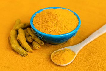 Turmeric powder in blue bowl with wooden spoon and dried roots at orange background.