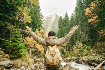 A man in a winter jacket in an evergreen forest against the backdrop of mountains and fog raised his hands up feeling the freedom and greatness of nature.