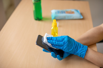 Woman's hand in blue gloves sanitizing cleaning smartphone mobile phone with alcohol on wood table surface with wet wipes