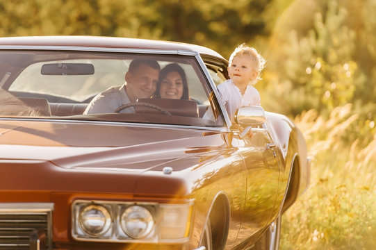 Buick Riviera In Retro Style On Sunset. Unique Car. Cute Blond Boy Is Sitting Behind The Wheel Of A Retro Car With His Family. Parents Are Sitting In The Back Seat, Boy Peeps Out The Window.