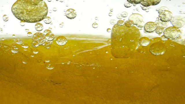 Golden liquid is stirred in a transparent bowl on a white background. Black cumin oil does not mix with water, close-up view. Sunflower oil gurgles in water and creates a bubble structure of air.