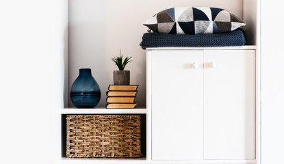 Cozy home interior decor: pillow, plaid, blue vase, stack of books, plant in pot, wicker box on a white shelf in the room. The quarantine concept of stay home, distance home education background.