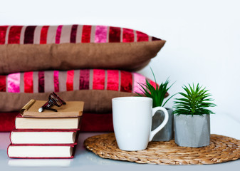 Cozy home interior decor: cup of coffee, stack of books, plants in pots on a wicker stand, pillows and plaid on a white table. Distance home education. Quarantine concept of stay home.