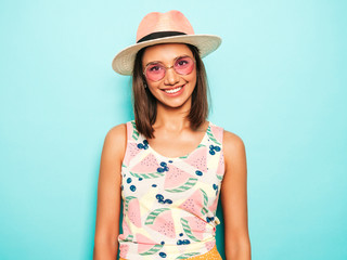 Young beautiful woman looking at camera in hat.Trendy girl in casual summer white T-shirt and yellow skirt in round sunglasses. Positive female shows facial emotions. Funny model isolated on blue