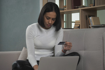 Black afro female using debit card shopping with laptop in living room. African American female entering credentials on laptop while sitting on soft sofa and shopping online