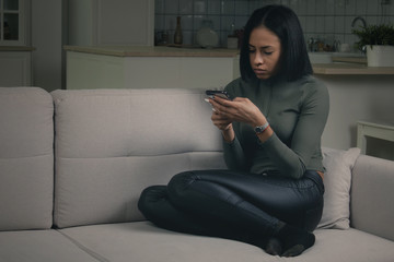 Beautiful young African American female in casual clothes browsing smartphone while resting on cozy sofa at home