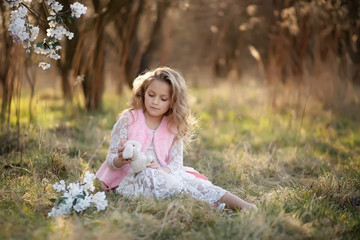 fair-haired girl of 7 years old, sitting in the field. holding a soft toy in his hands. dressed in a long lace white dress and a pink vest. field in the background