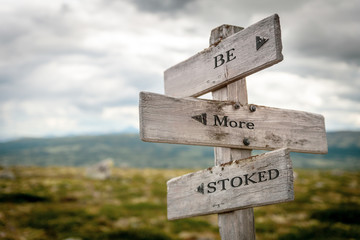 be more stoked text engraved on old wooden signpost outdoors in nature. Quotes, words and illustration concept.