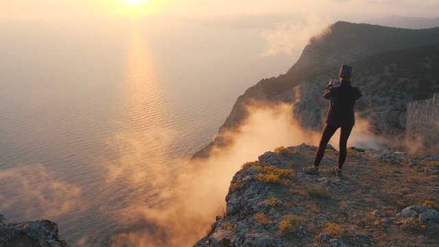 Young woman tourist stands at a cliff and shoots a video on a smartphone. Unusual steam, evaporation and smoke from clouds under the mountain against the backdrop of sunset over the sea in autumn.
