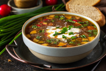 Traditional dish Russian cuisine. Cold summer Okroshka soup close-up with kvass, sausage and vegetables in a bowl on a dark stone countertop.