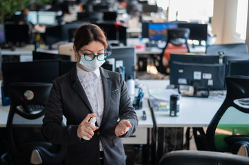 Female manager in a medical mask sprays on hand antiseptic. A woman in a suit uses a sanitizer to disinfect. Concept of office work during the coronavirus epidemic.