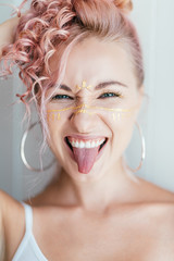 Be cheerful. Portrait of young beautiful woman with pink hair and artistic makeup smiling at camera, sticking her tongue out, posing over light white background. Face art concept. Vertical shot