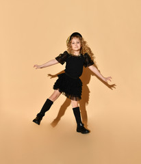 Fototapeta na wymiar Charming girl with blond hair in black dress and golden cap stands on one leg looking down on her stylish black boots, isolated on peach background