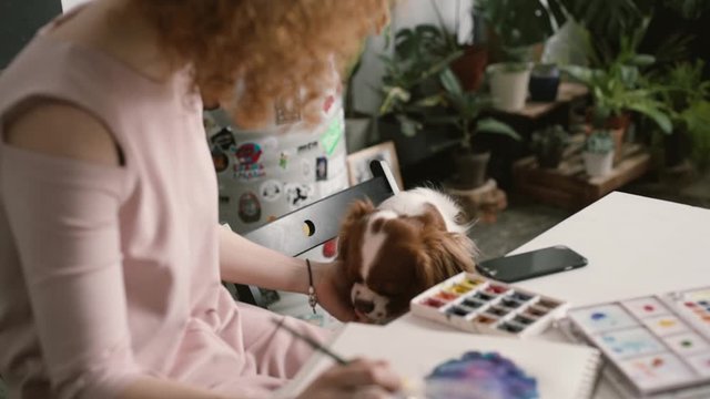 red-haired girl paints with watercolors and a dog runs up to her
