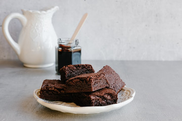 Homemade dark chocolate brownies and sauce in glass jar on gray background. 