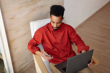 Top view of young brunette dark skinned man dressed in red shirt while sitting in chair over beige interior with laptop and checking messages on his phone