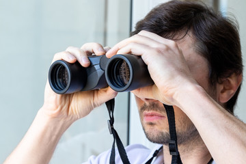 Fototapeta na wymiar Handsome bearded man looking through binoculars at the window with a view of the neighboring house for observing neighbors, environment or nature during quarantine. Selective focus. Closeup view