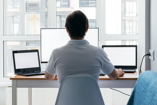 A man is sitting at a workplace with two laptops and a monitor near the window. Remote work from home in quarantine. Back view
