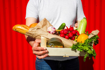 Box with vegetables, fruits, bread and canned goods in male hands on a red background.