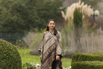 Fototapeta premium Beautiful Indian woman wearing poncho posing in ornamental garden. Looking away.