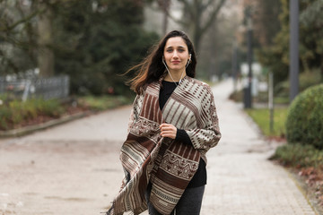 Young Indian woman dressed in poncho and listening to earphones walking towards the camera.