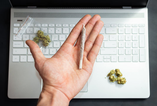 Top View Of Hand Holding A Big Marijuana Joint With Computer And Cannabis Buds On Background. Concept Of Marijuana And Technology Or Office.