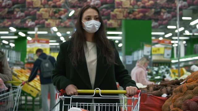 Young Woman Wearing Protective Medical Face Mask Rolls Shopping Cart At Grocery Store. Female Customer Picks Vegetables At Economy Supermarket During Covid-19 Coronavirus Pandemic, Social Distancing