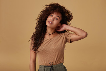 Attractive young dark skinned brunette woman with wild curly hair raising hand to her head and looking calmly at camera, standing over beige background