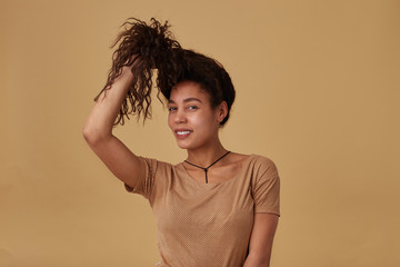 Positive young lovely brown haired curly female with dark pulling her hair with raised hand and smiling cheerfully at camera, isolated over beige background