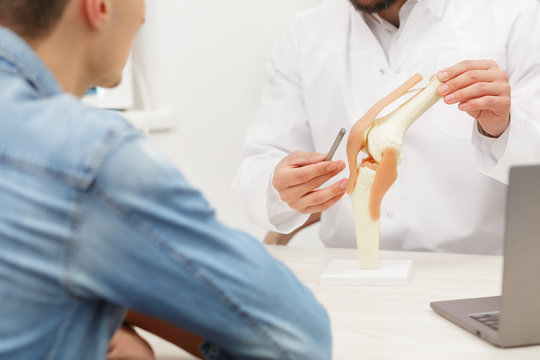 Doctor showing flexible knee joint model to his patient and explains the cause of his pain in medical office. Patient visiting physiotherapist in clinic. Surgery, healthcare and medicine concept.