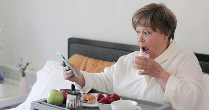 Side view of old beautiful lady making amazed facial expession while using her modern mobilephone. Elderly woman having breakfast and drinking juice while getting shocking news.
